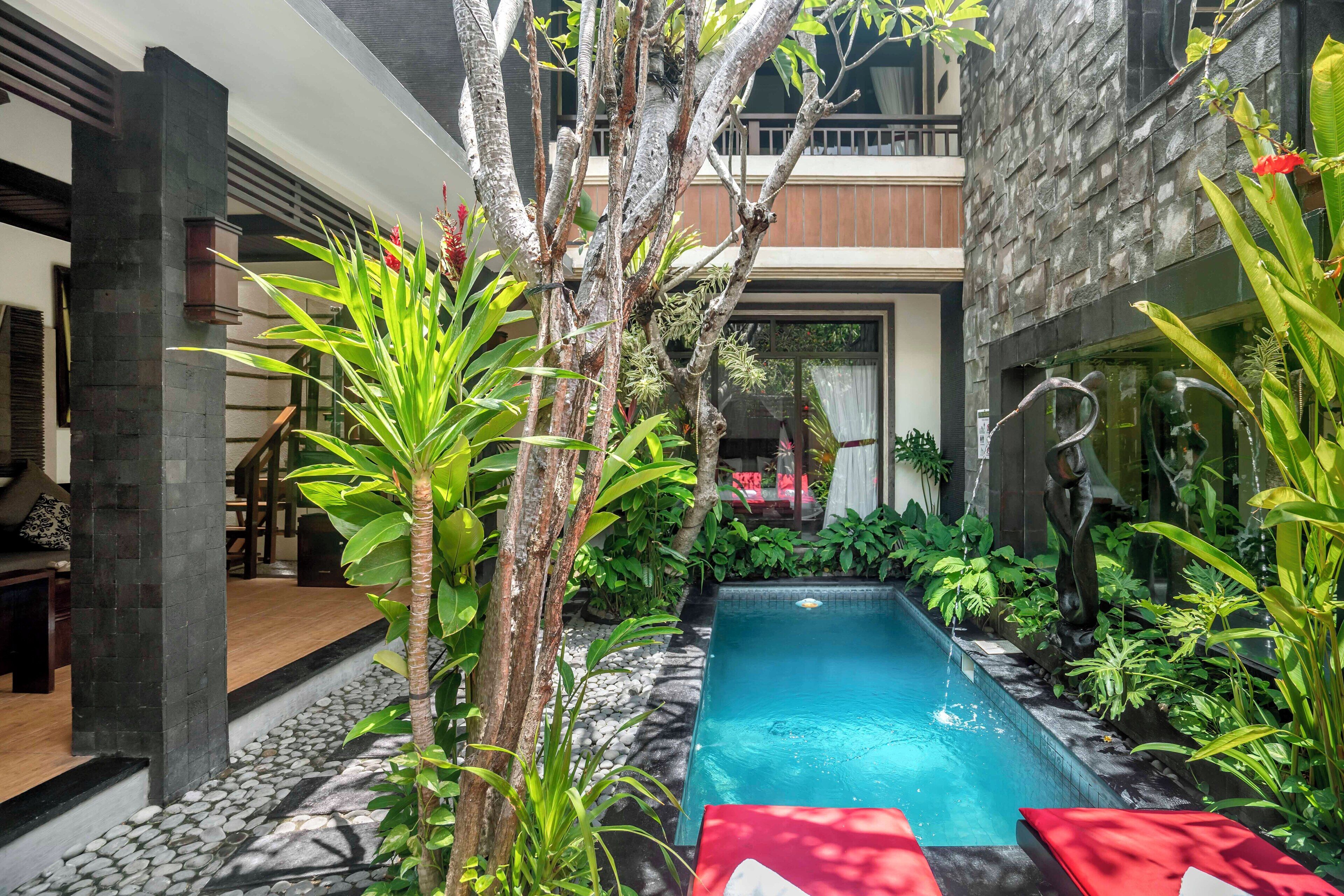 A pool area with a stone pathway, surrounded by lush greenery and a modern building with a balcony.