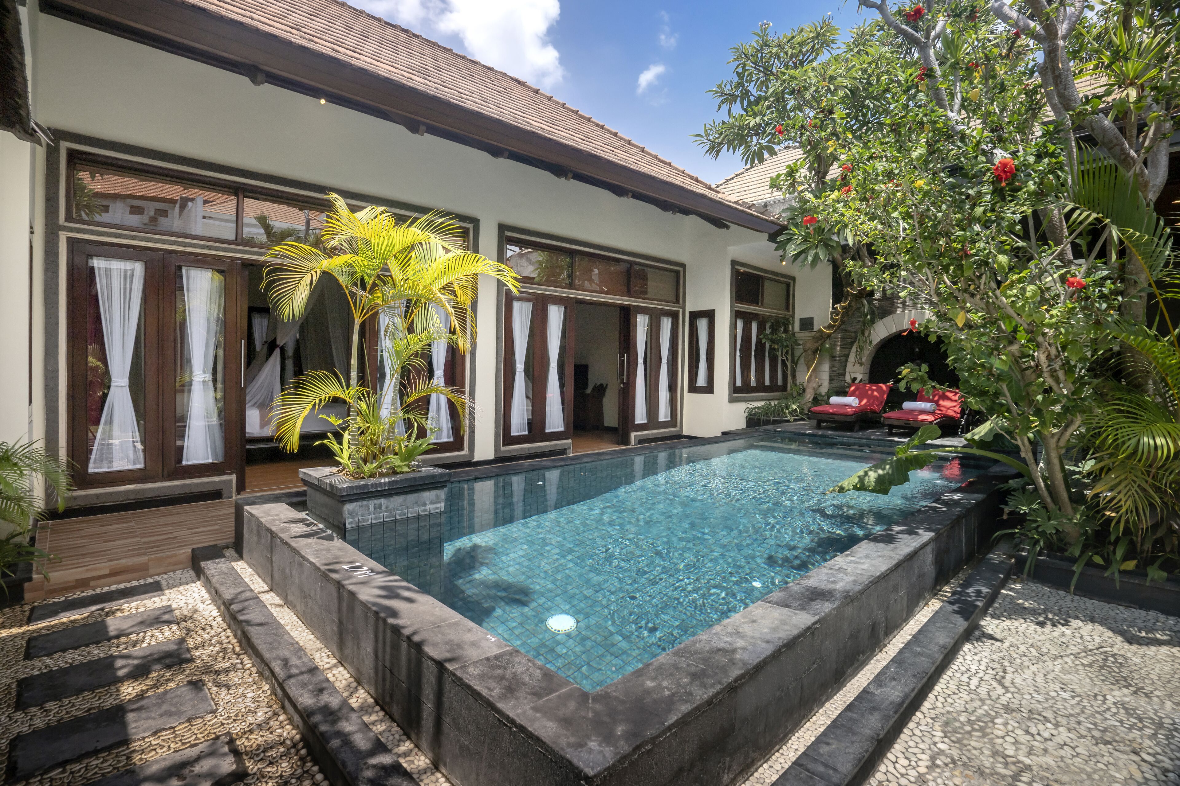 A swimming pool with a tiled deck, surrounded by a garden with tropical plants and a red chair.