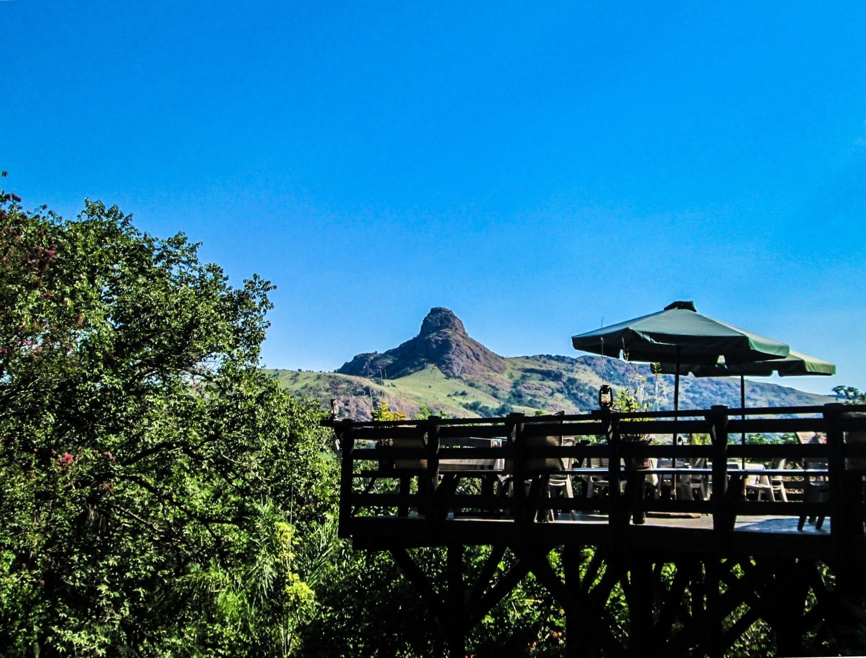 Una terraza de madera con mesas y sillas, con vistas a una montaña y vegetación exuberante.
