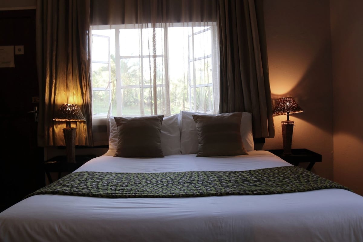 Una habitación de hotel con una cama grande, dos lámparas de noche, un ventanal con cortinas y vistas al exterior con vegetación.