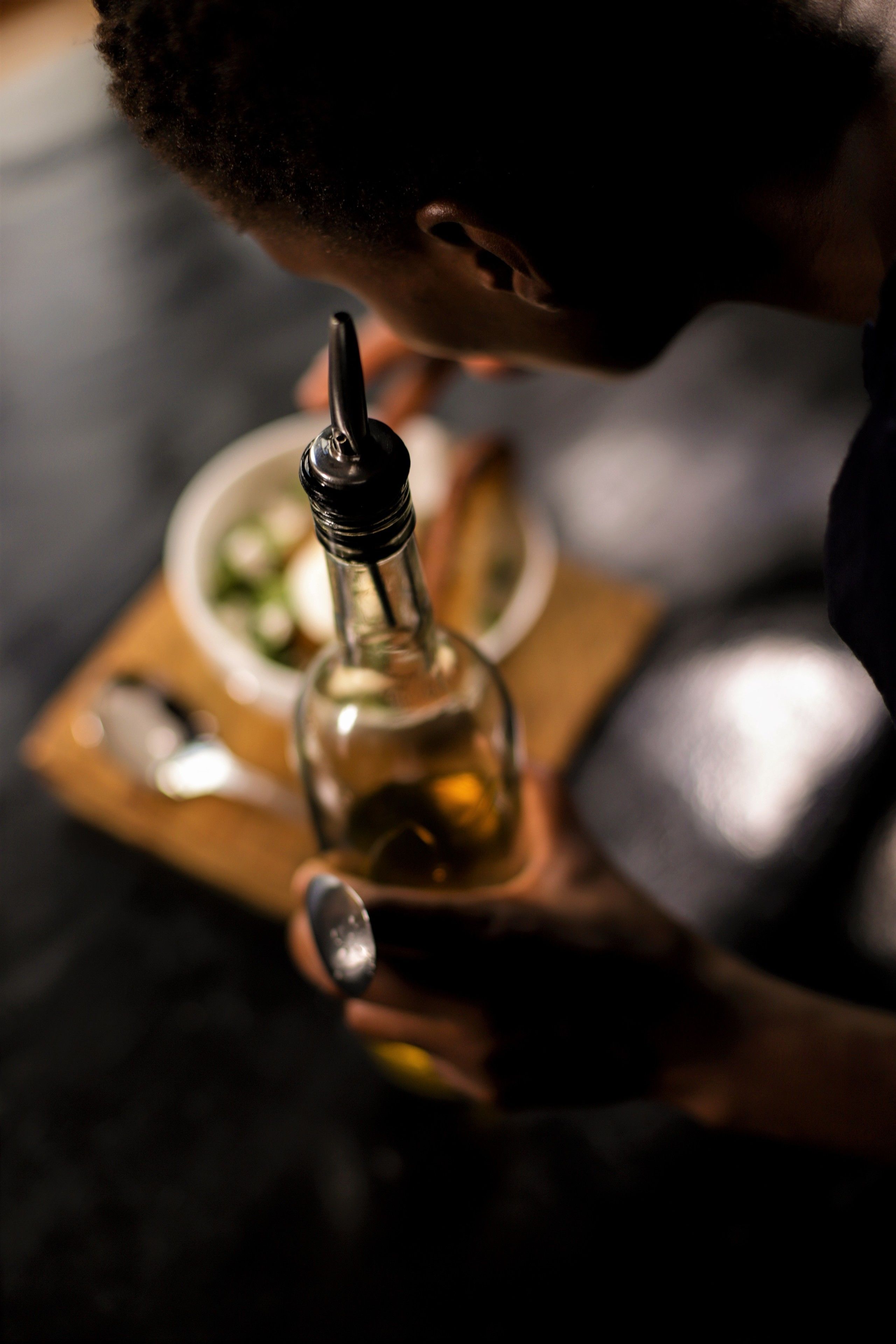 A person holding a bottle of olive oil with a dropper, a bowl of salad, and a spoon on a wooden board.