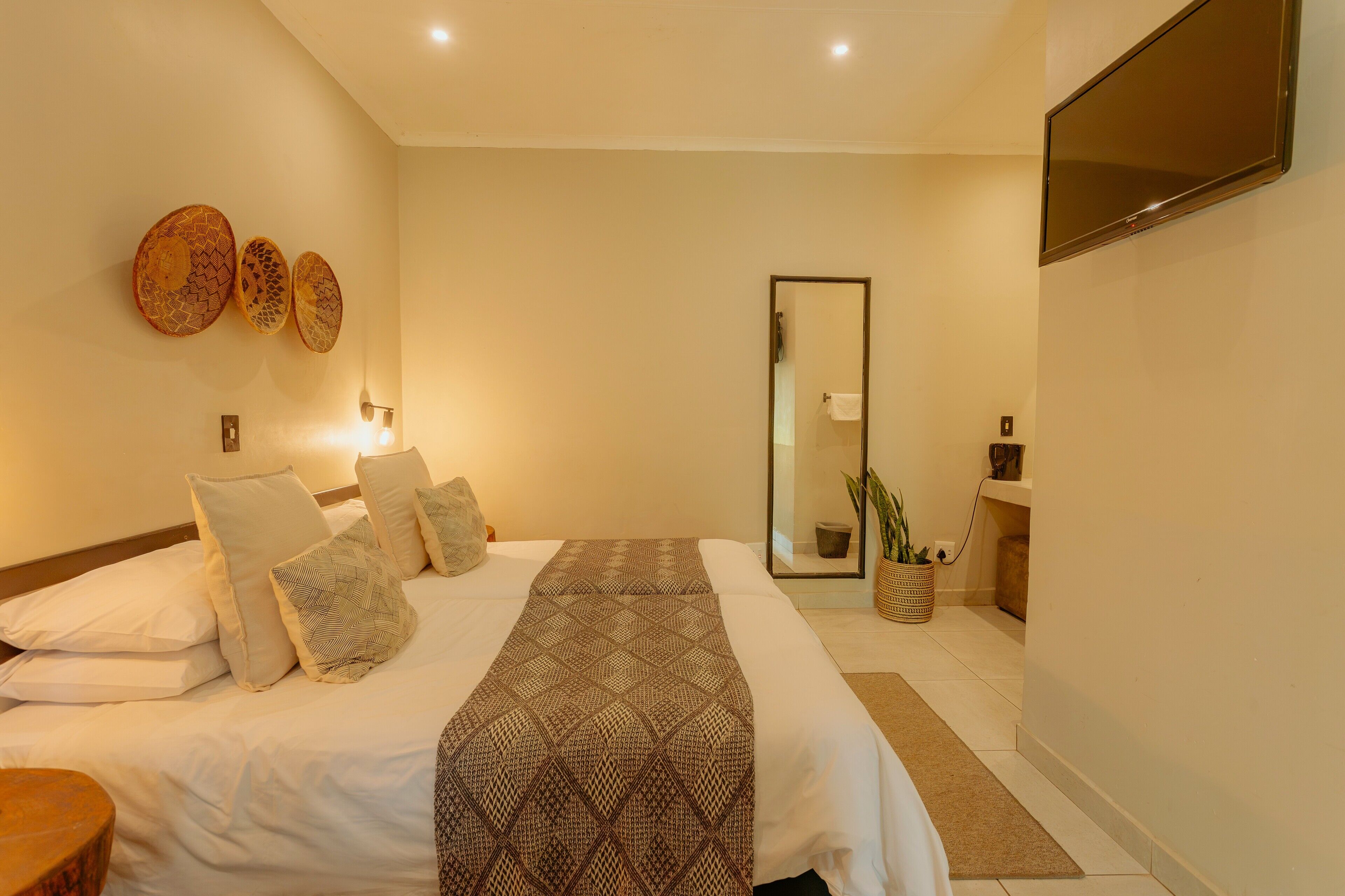 A modern bedroom with a large bed, a flat-screen TV, a mirror, and a small plant.