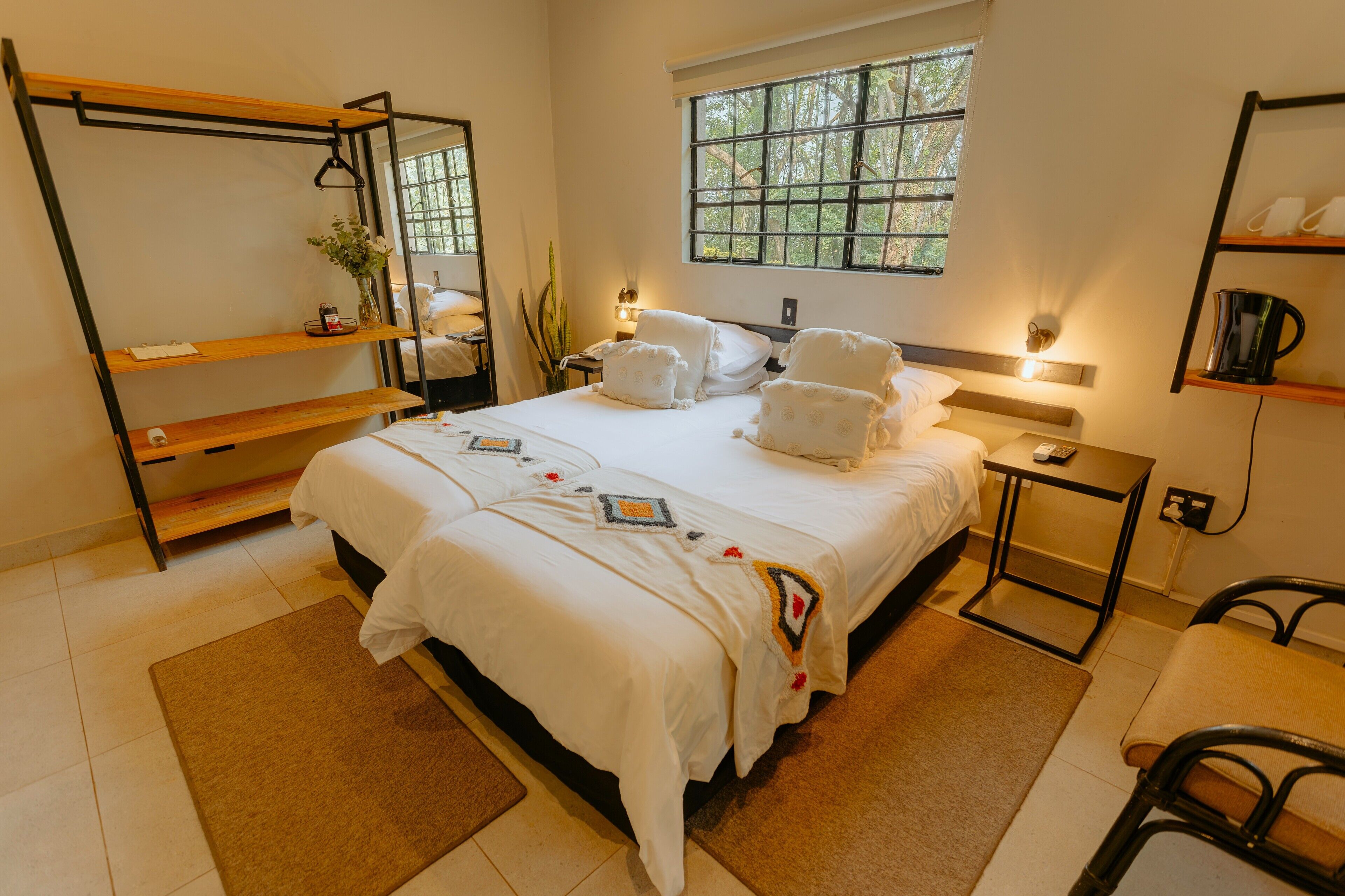 A hotel room with a large bed, a bedside table, a chair, a window with a view of trees, and a shelf with towels.