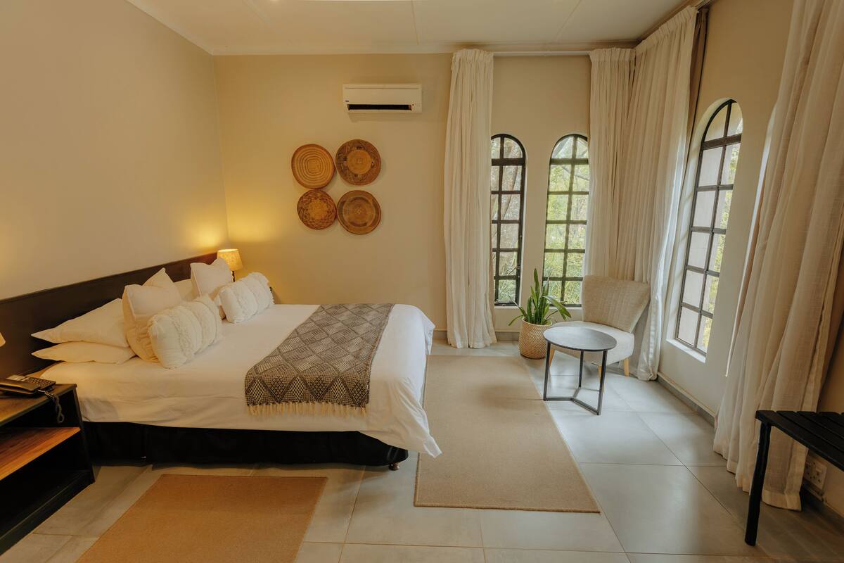 Habitación de hotel con cama, mesita de noche, silla y ventanas amplias con cortinas.