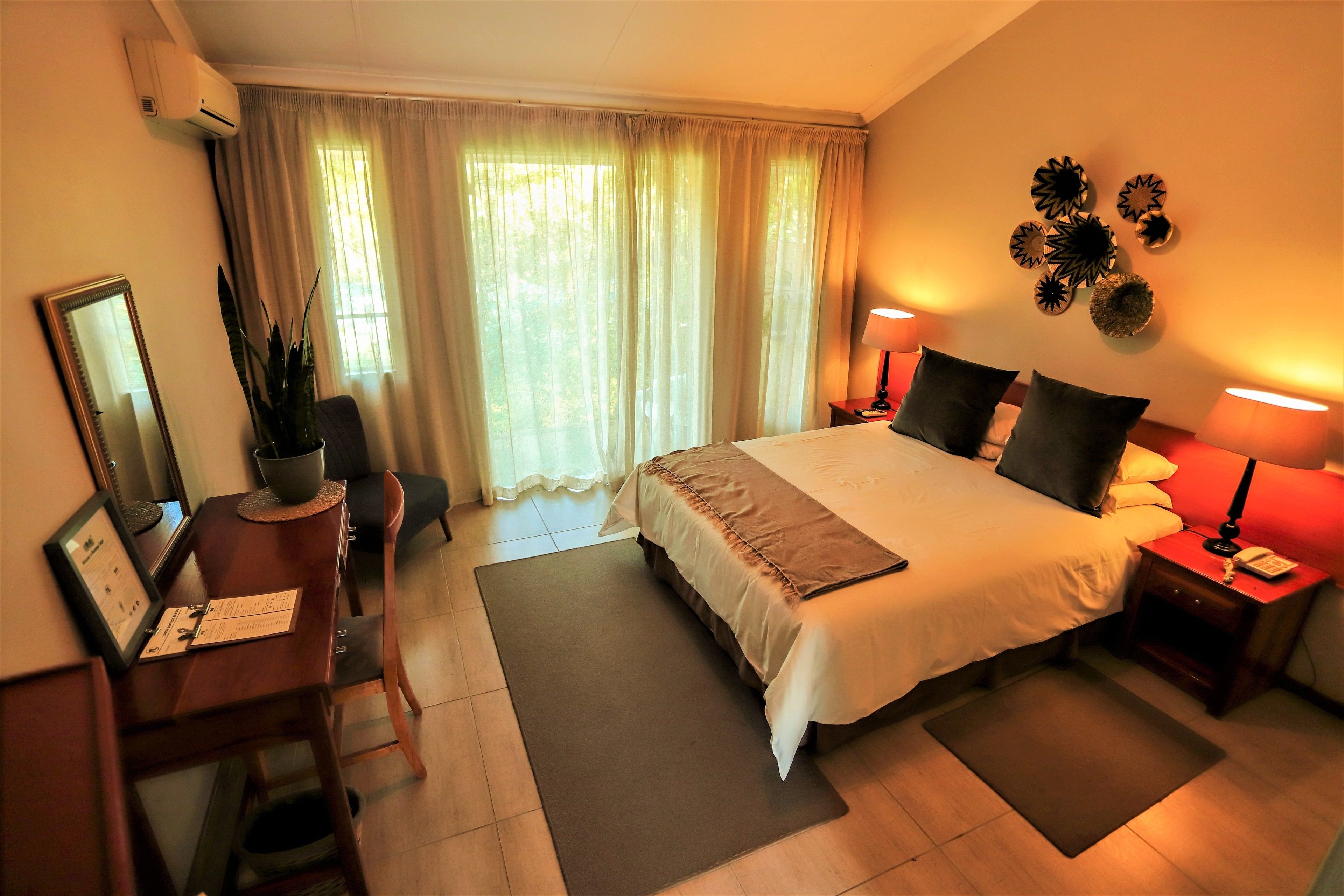 A hotel room with a large bed, bedside lamps, a desk with a chair, a mirror, and a window with curtains.