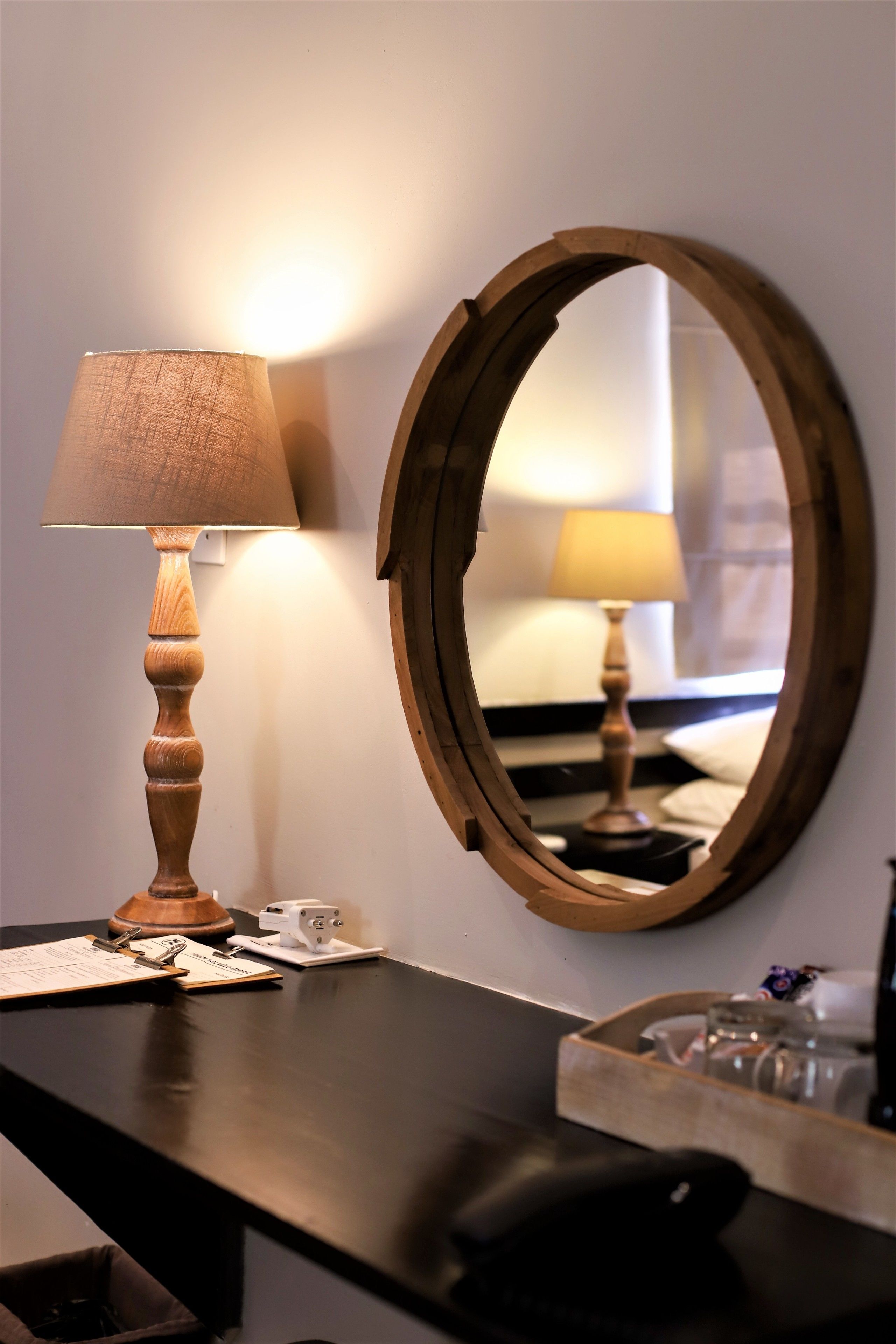 A wooden-framed mirror on a wall, a wooden lamp with a fabric shade, and a black table with a tray of glasses.