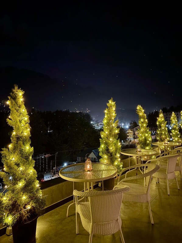 Una terraza en la azotea con mesas redondas y sillas de mimbre, decorada con árboles de Navidad y luces colgantes.