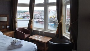 Habitación de hotel con cama, una silla, una mesita con botellas de agua y vista a un puerto con barcos y edificios.