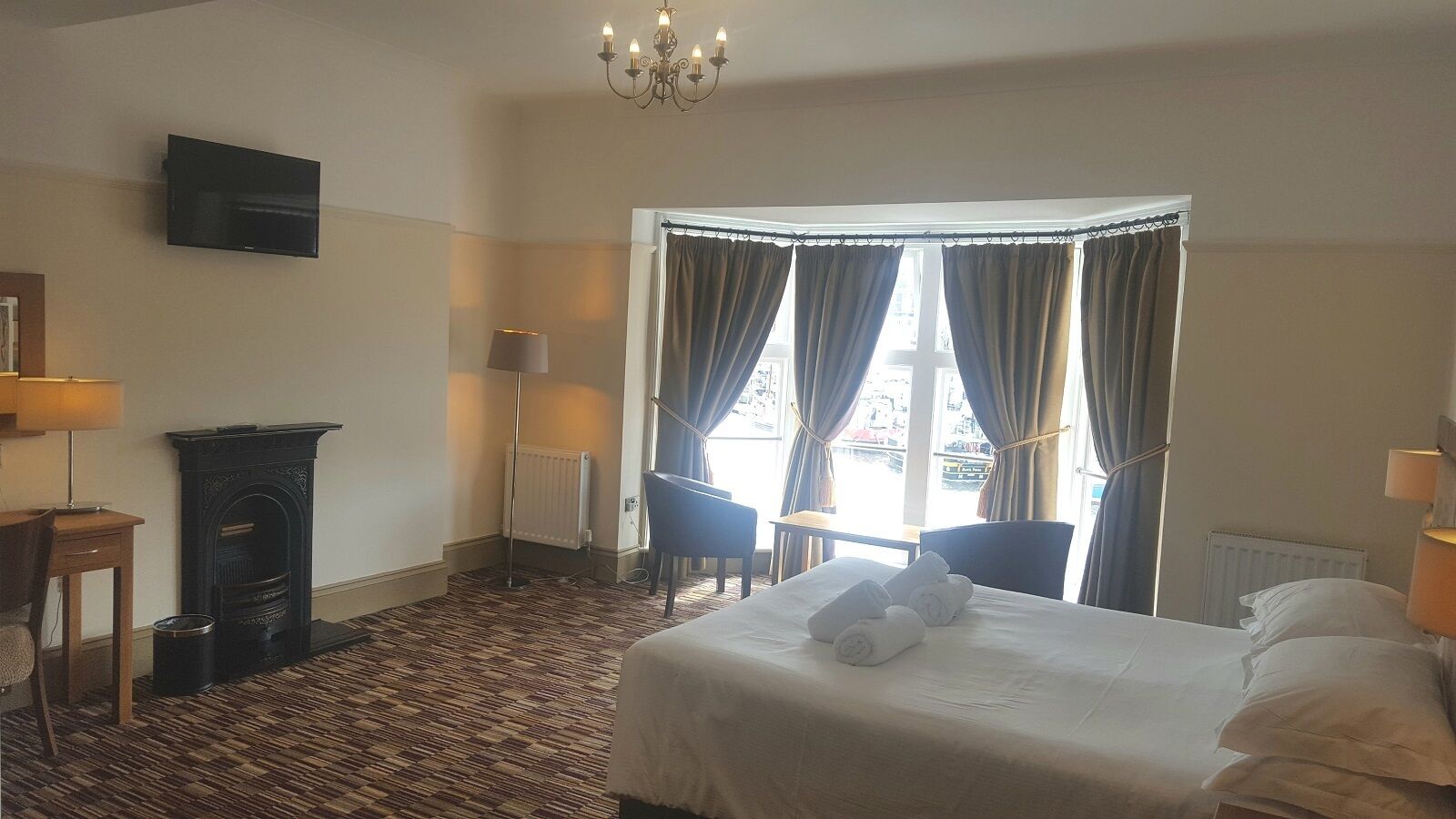 A hotel room with a bed, two chairs, a fireplace, a TV, and a window with curtains.