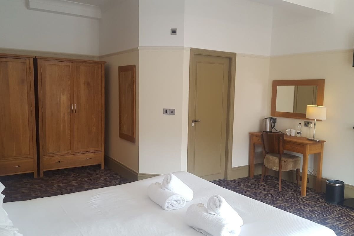 Une chambre d’hôtel avec un grand lit, une armoire en bois, un bureau avec une chaise, un miroir et une cheminée.