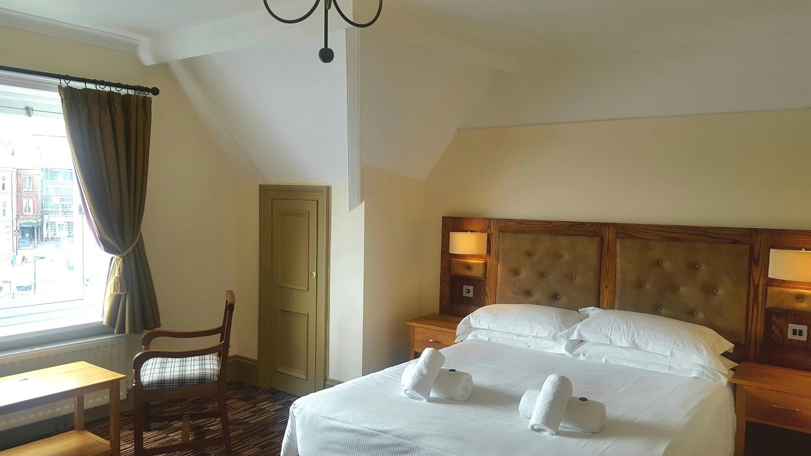 A hotel room with a large bed, a wooden headboard, a nightstand, a chair, and a window with curtains.