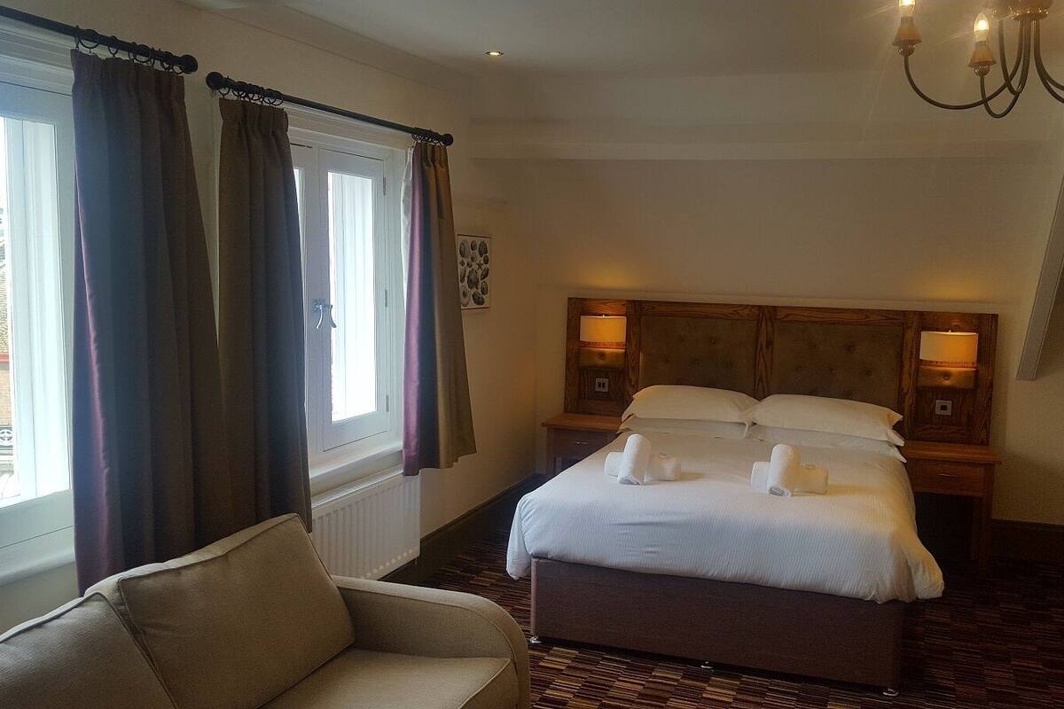 Une chambre d’hôtel avec un lit, deux tables de chevet avec des lampes, une chaise et une fenêtre avec des rideaux.