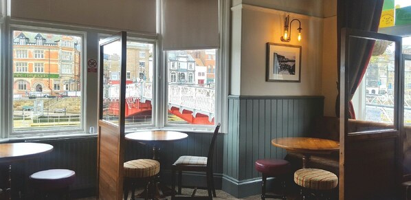 A corner seating area with two tables and chairs, large windows with a view of buildings, and a framed picture on the wall. - The Dolphin (Whitby)