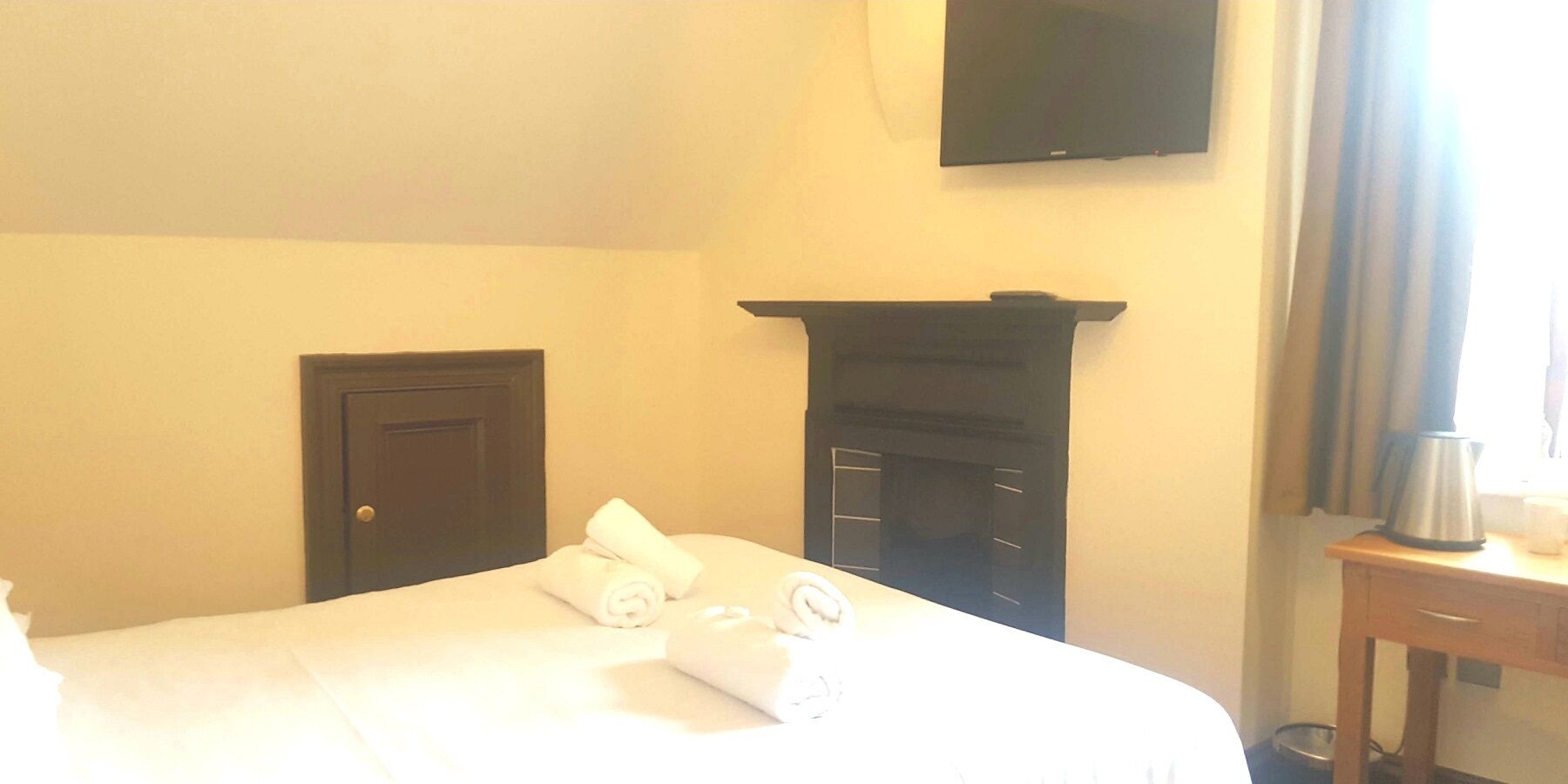 A hotel room with a bed, a flat-screen TV mounted on the wall, a fireplace, and a small table with a kettle.