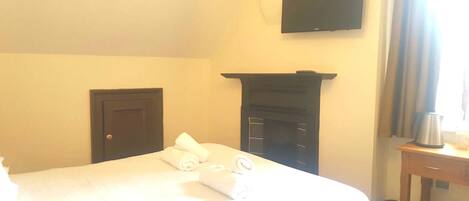 A hotel room with a bed, a flat-screen TV mounted on the wall, a fireplace, and a small table with a kettle.