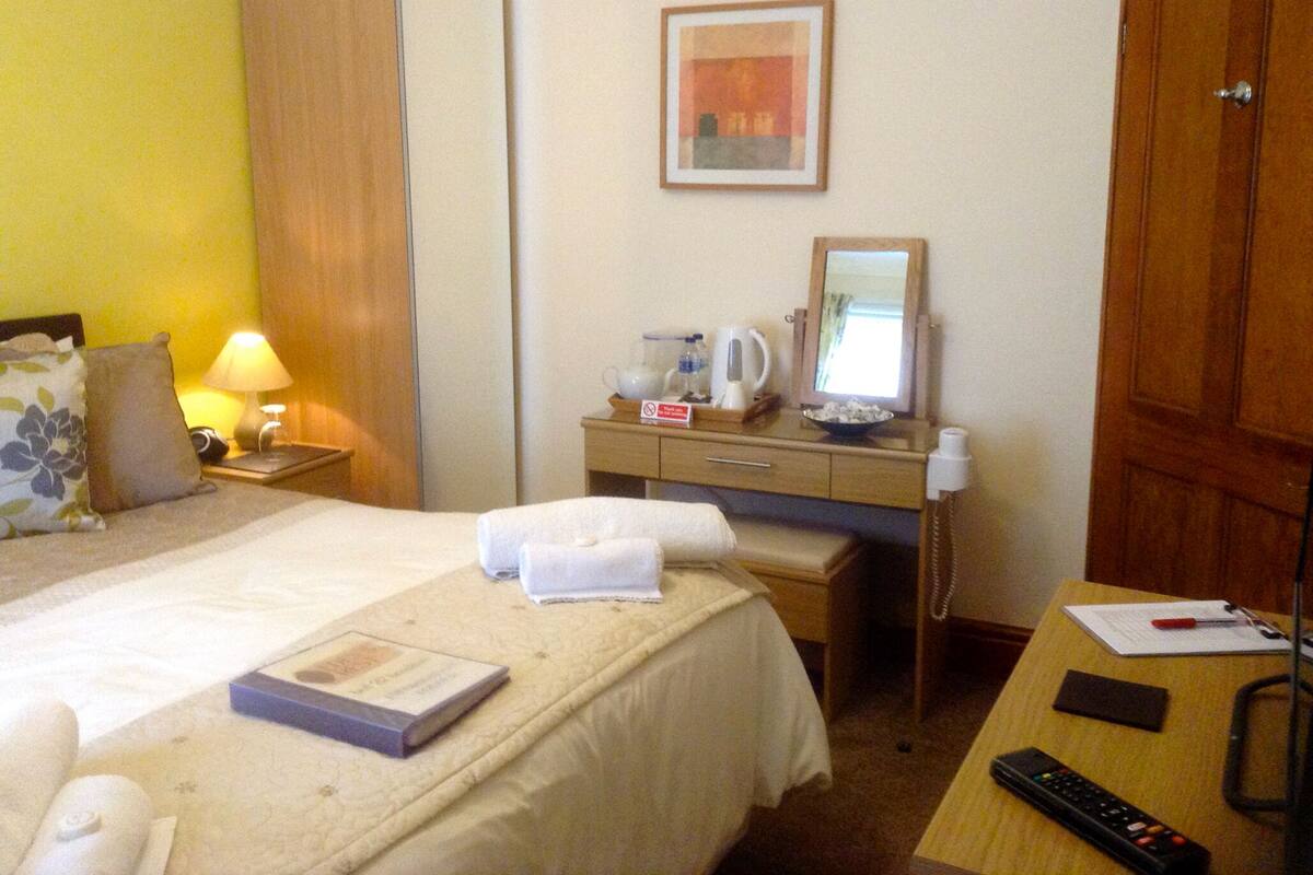Une chambre d’hôtel comprenant un lit, un bureau, une armoire, une table de chevet avec une lampe, une machine à café, un bol, un miroir et une télécommande.