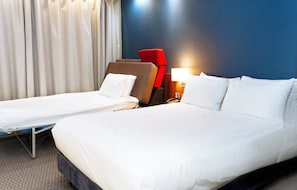 A hotel room with a large bed, two single beds, a red chair, a lamp, and a curtain. - Holiday Inn Express Harlow by IHG (Harlow)