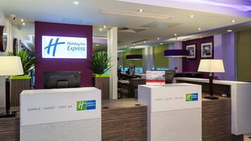 Zona de recepción de Holiday Inn Express con un diseño moderno, que incluye un mostrador de recepción, una pantalla grande con el nombre del hotel y un cartel que da la bienvenida a los miembros de IHG Rewards Club.