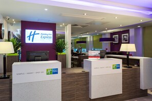 A Holiday Inn Express reception area with a modern design, including a reception desk, a large screen displaying the hotel name, and a sign welcoming IHG Rewards Club members. - Holiday Inn Express Harlow by IHG (Harlow)