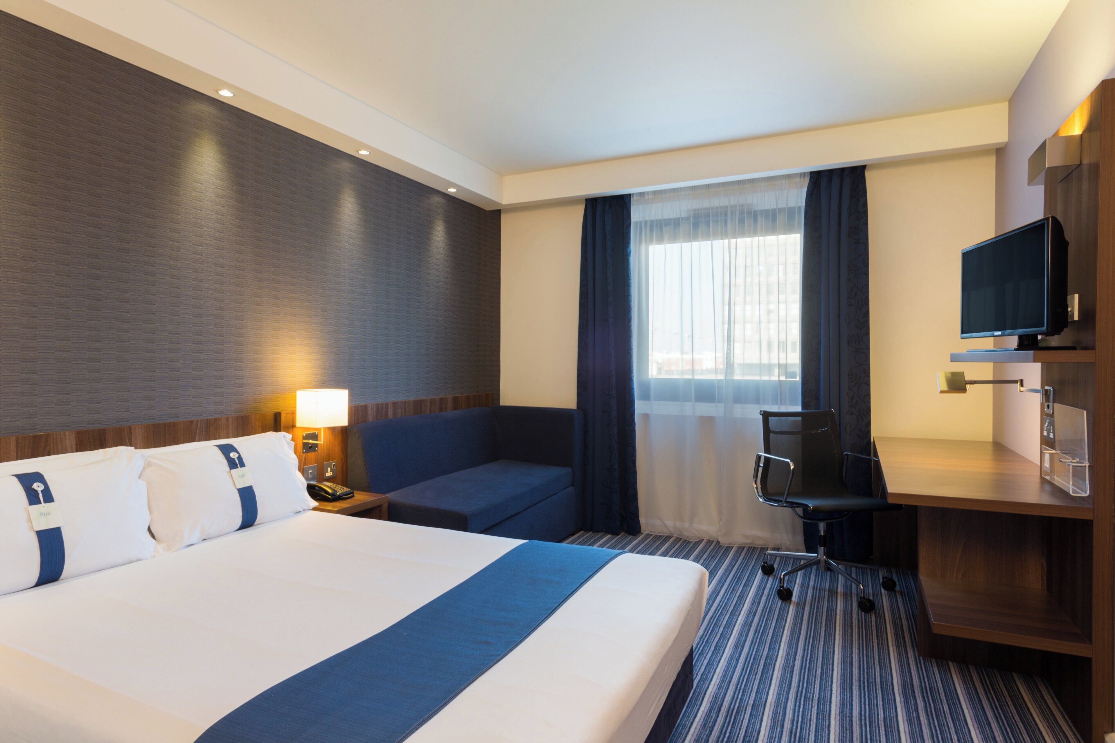 A hotel room with a large bed, a desk, a chair, a television, and a window with curtains.