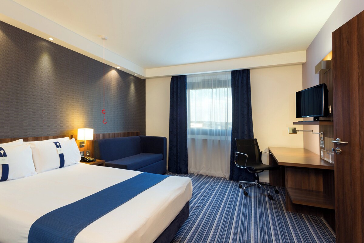 A hotel room with a large bed, a desk with a chair, a television, and a window with curtains.