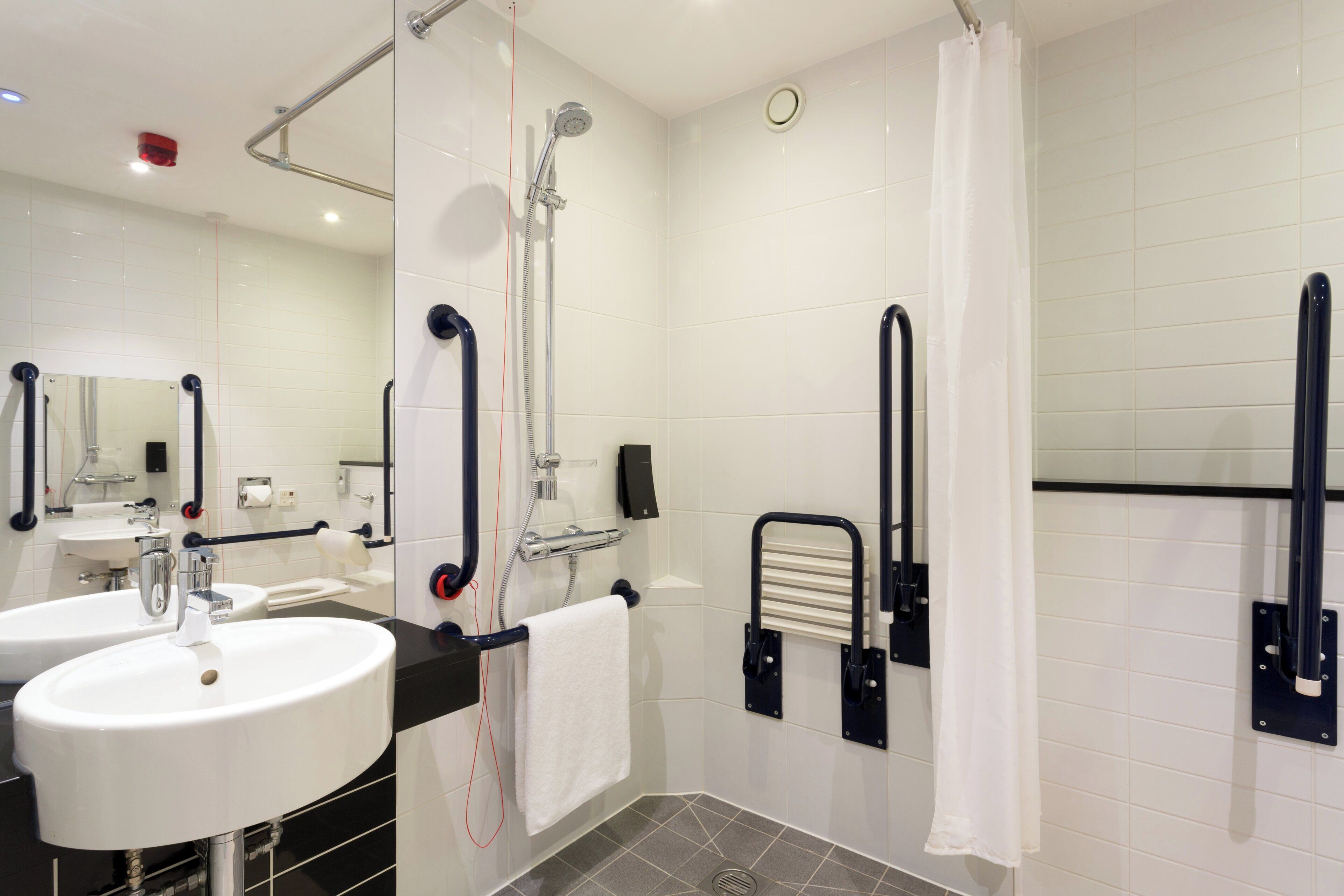 A modern bathroom with two sinks, a shower with a handheld showerhead, a toilet with a grab bar, and a towel rack.