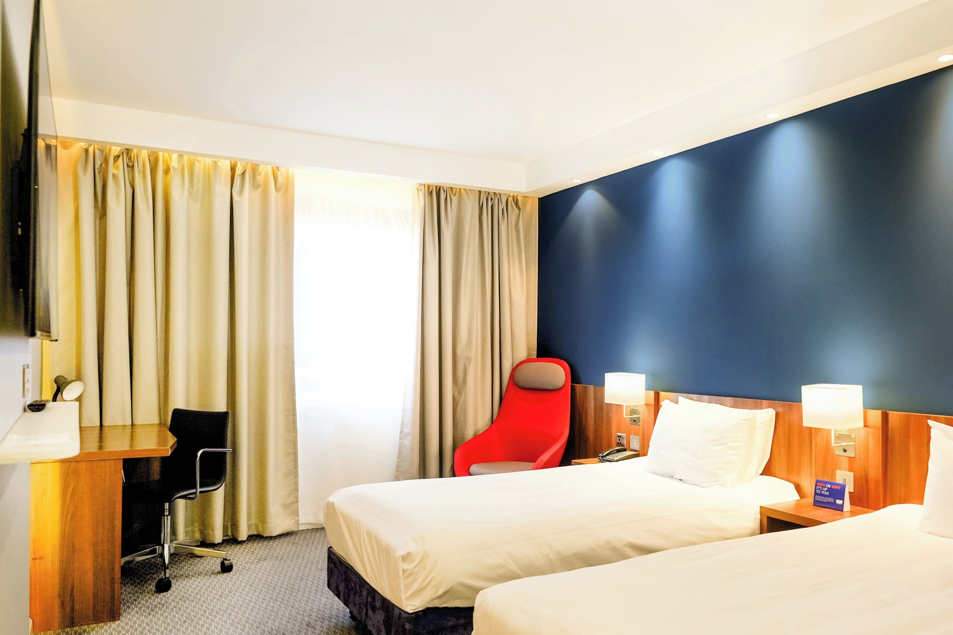 Ein Hotelzimmer mit zwei Betten, einem Schreibtisch, einem roten Sessel, einem Fernseher und einem Fenster mit Vorhängen.