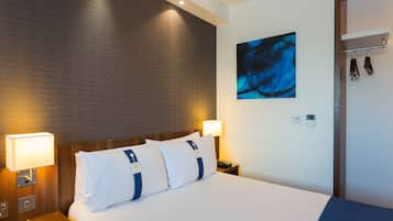 Una habitación de hotel con una cama bien hecha, dos lámparas de noche, una obra de arte en la pared y un perchero con objetos colgados.