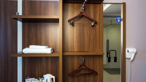 A wooden wardrobe with shelves holding towels and a kettle. A hairdryer is mounted on the wall next to a door. - Holiday Inn Express Harlow by IHG (Harlow)
