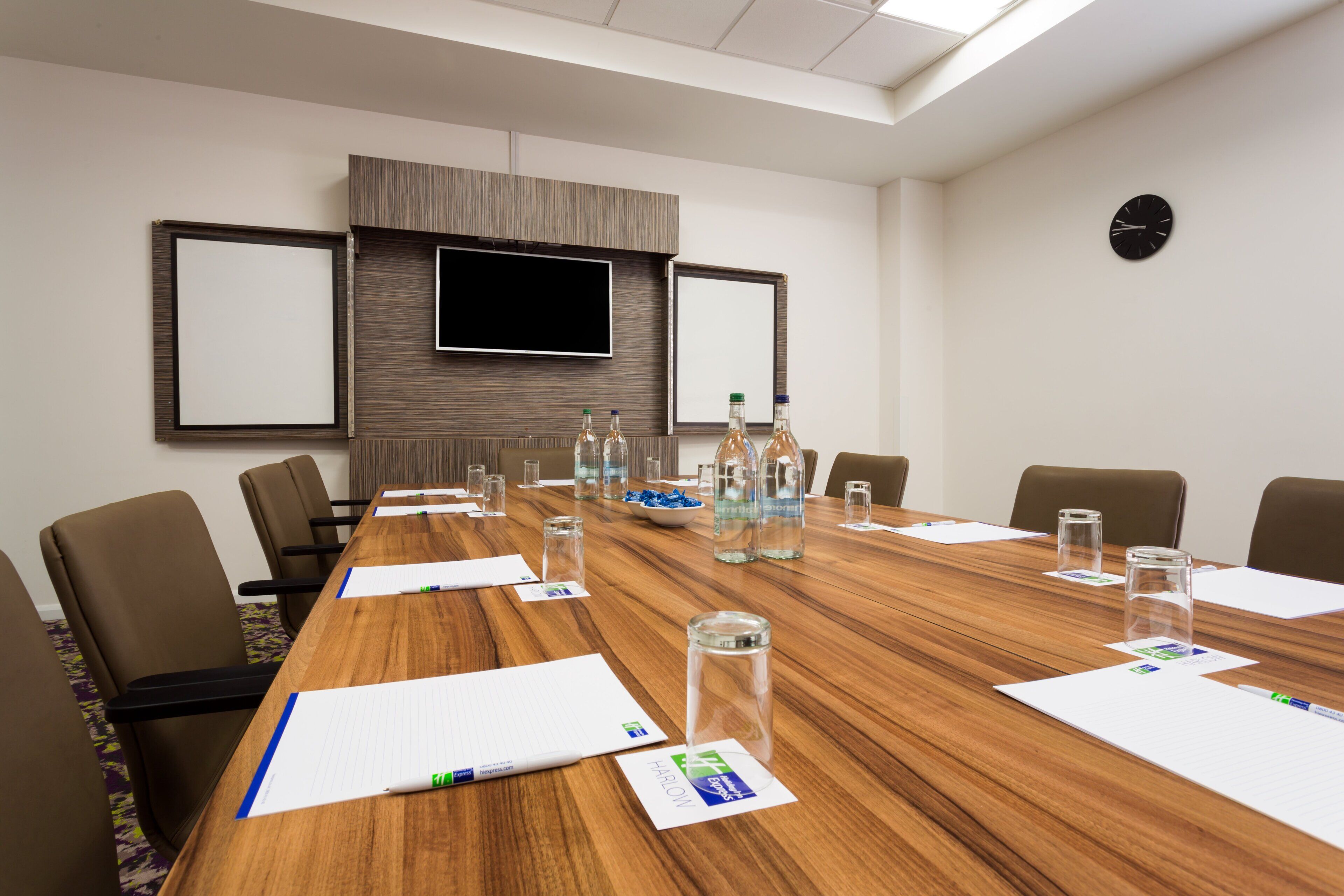 A conference room with a long wooden table, chairs, a flat-screen TV, and a clock on the wall.