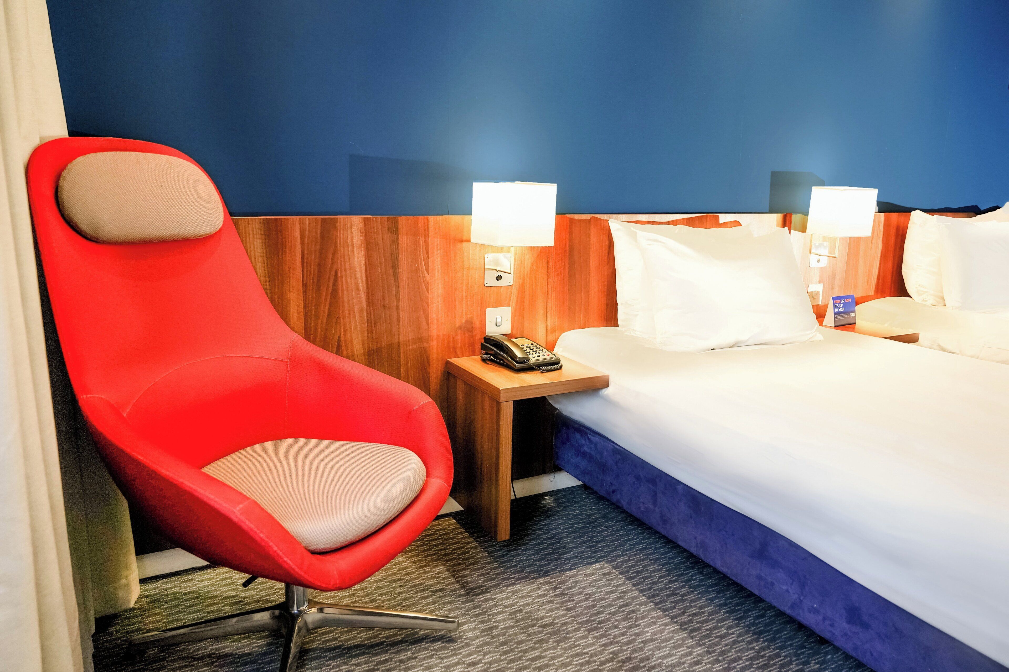 Une chambre d’hôtel avec un fauteuil rouge, un lit avec du linge de maison blanc, deux lampes de chevet et une tête de lit en bois.