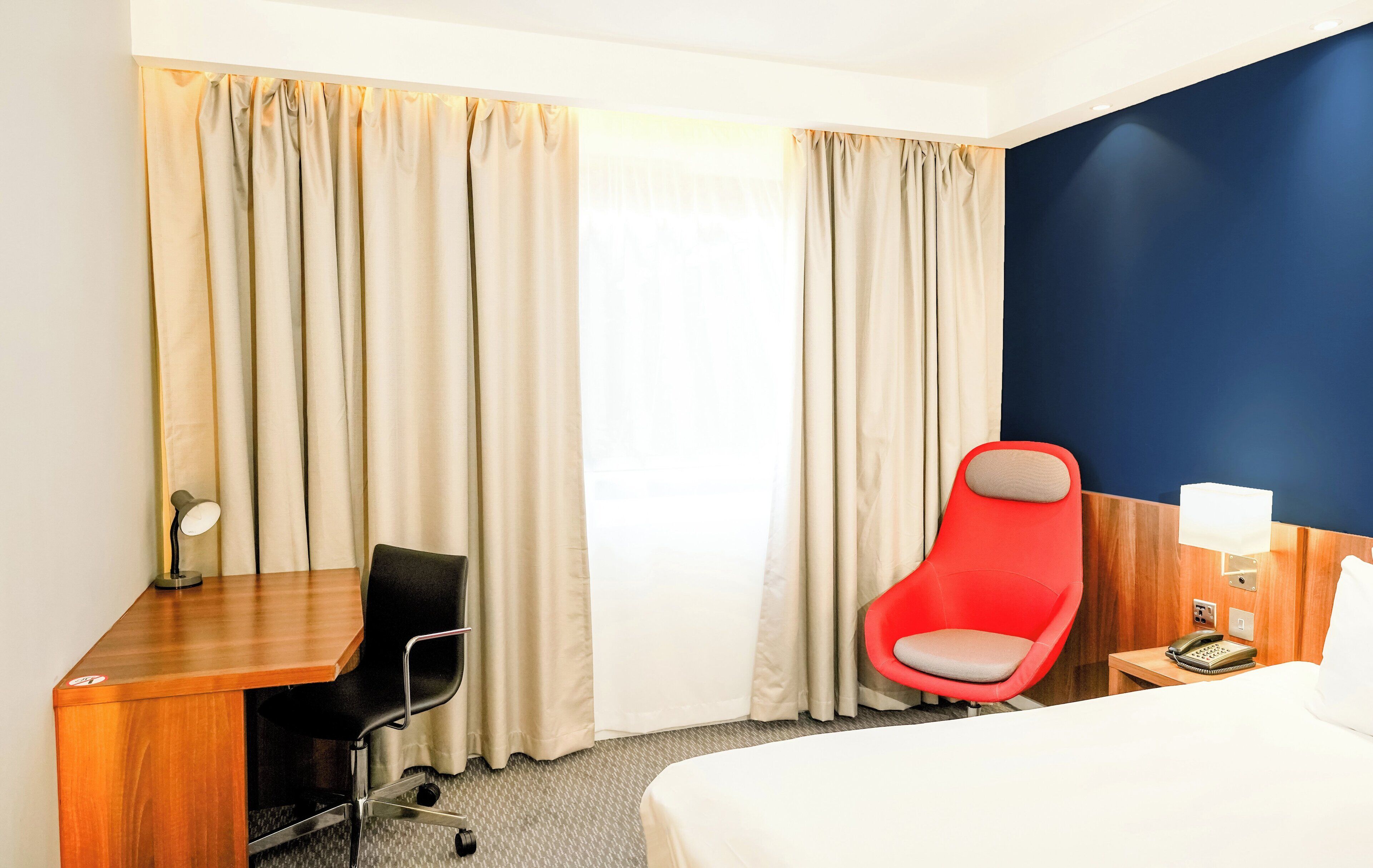 Una habitación de hotel con cama, escritorio y silla, una silla roja, mesita de noche con lámpara y cortinas.