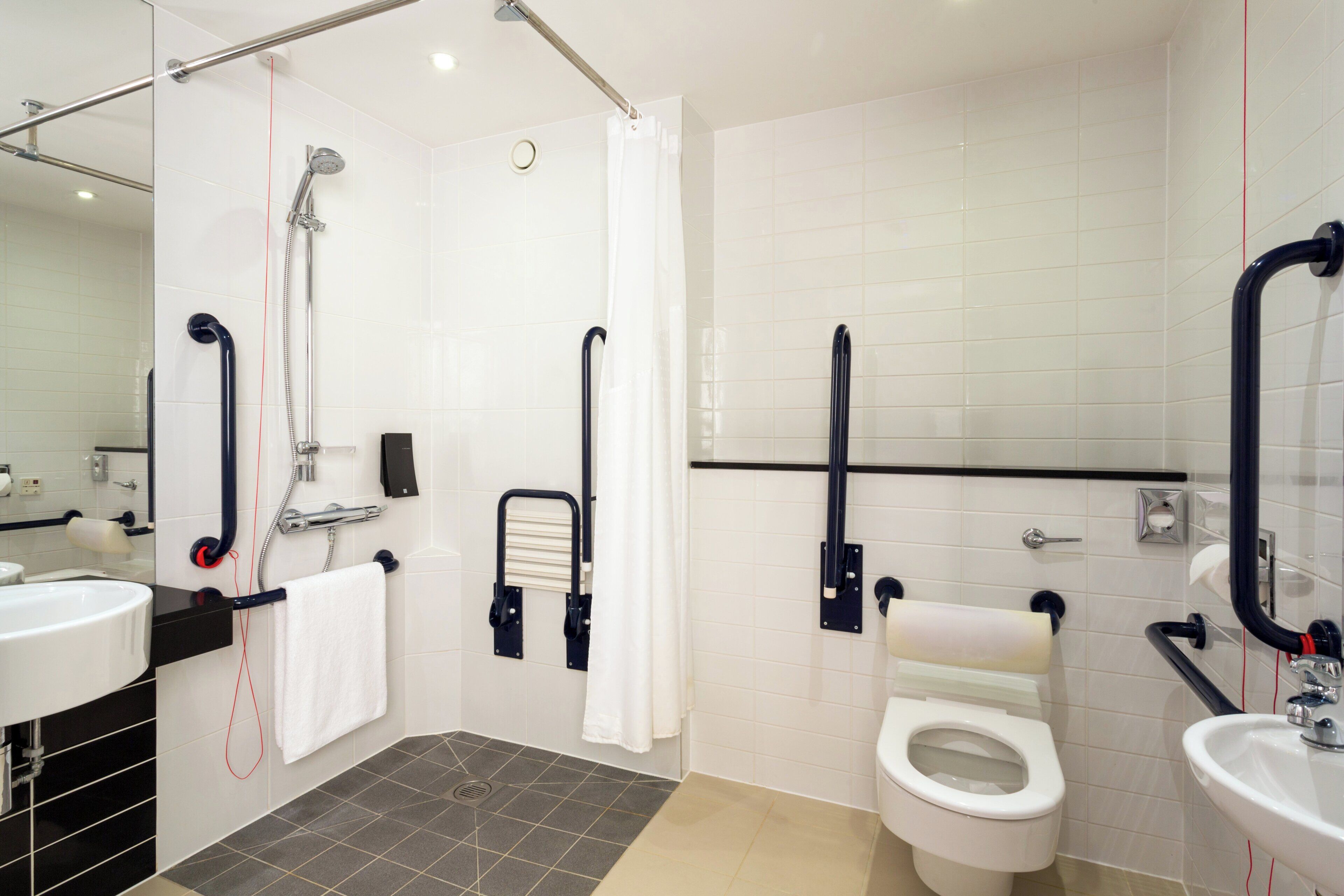 Une salle de bain moderne équipée d’une douche, de toilettes et d’un lavabo, et aménagée pour faciliter l’accès aux personnes à mobilité réduite, avec notamment des barres d’appui et une main courante.