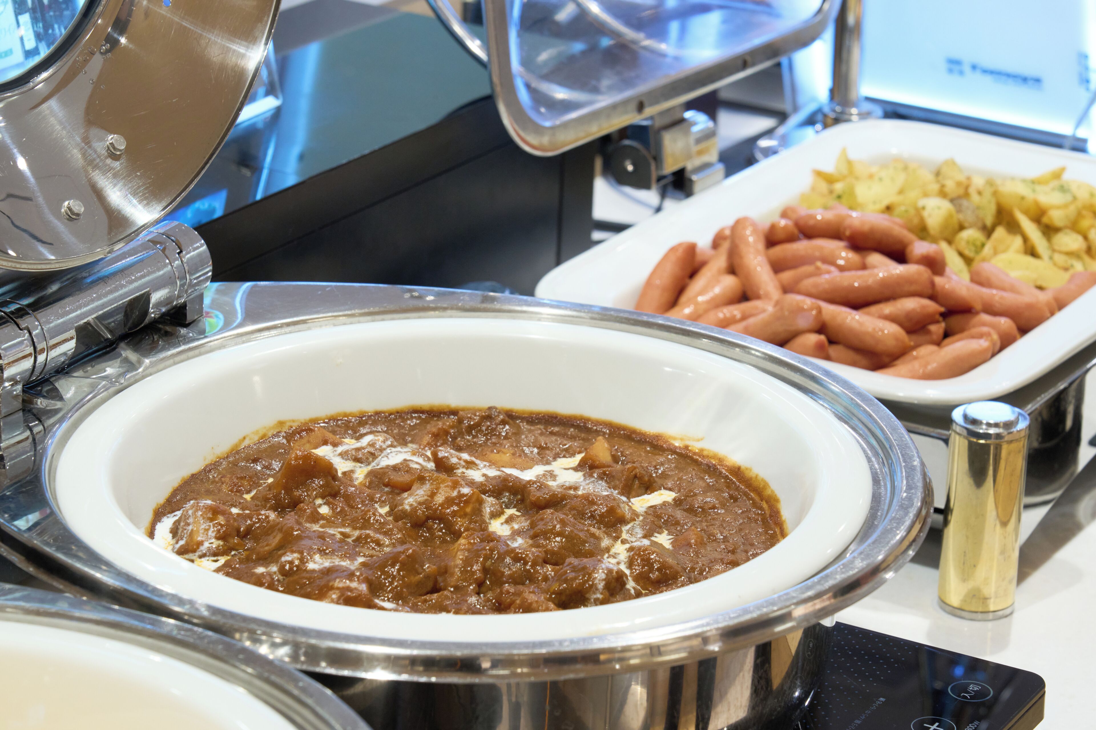 Ein reichhaltiges Buffet mit verschiedenen Speisen, darunter eine Schüssel Curry, Würstchen und Kartoffeln.