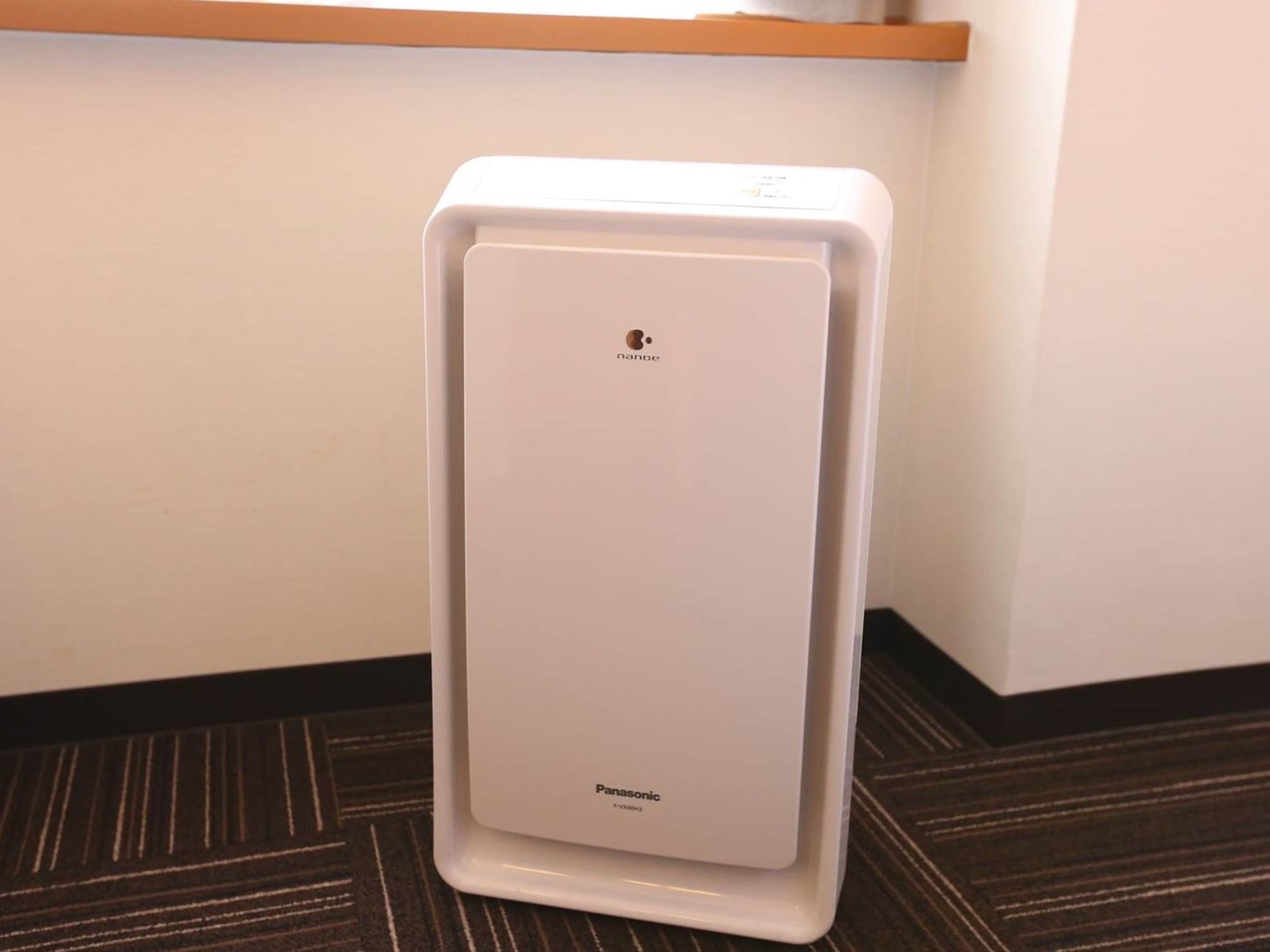 Ein weißes Panasonic Luftreinigergerät steht in einer Ecke eines Raumes mit dunklem Teppich und einem Holzboden darüber.