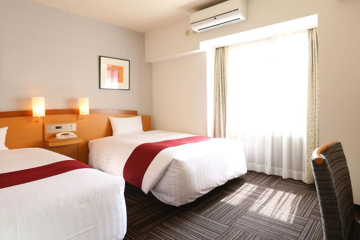 A hotel room with two beds, a bedside table with a phone, an air conditioner, and a large window with curtains.