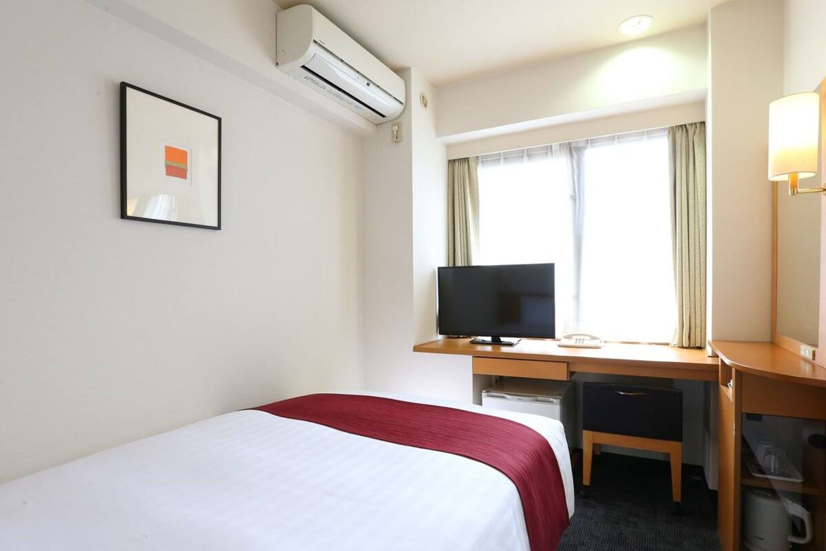 A hotel room with a bed, a desk with a computer, a chair, a desk lamp, and a window with curtains.