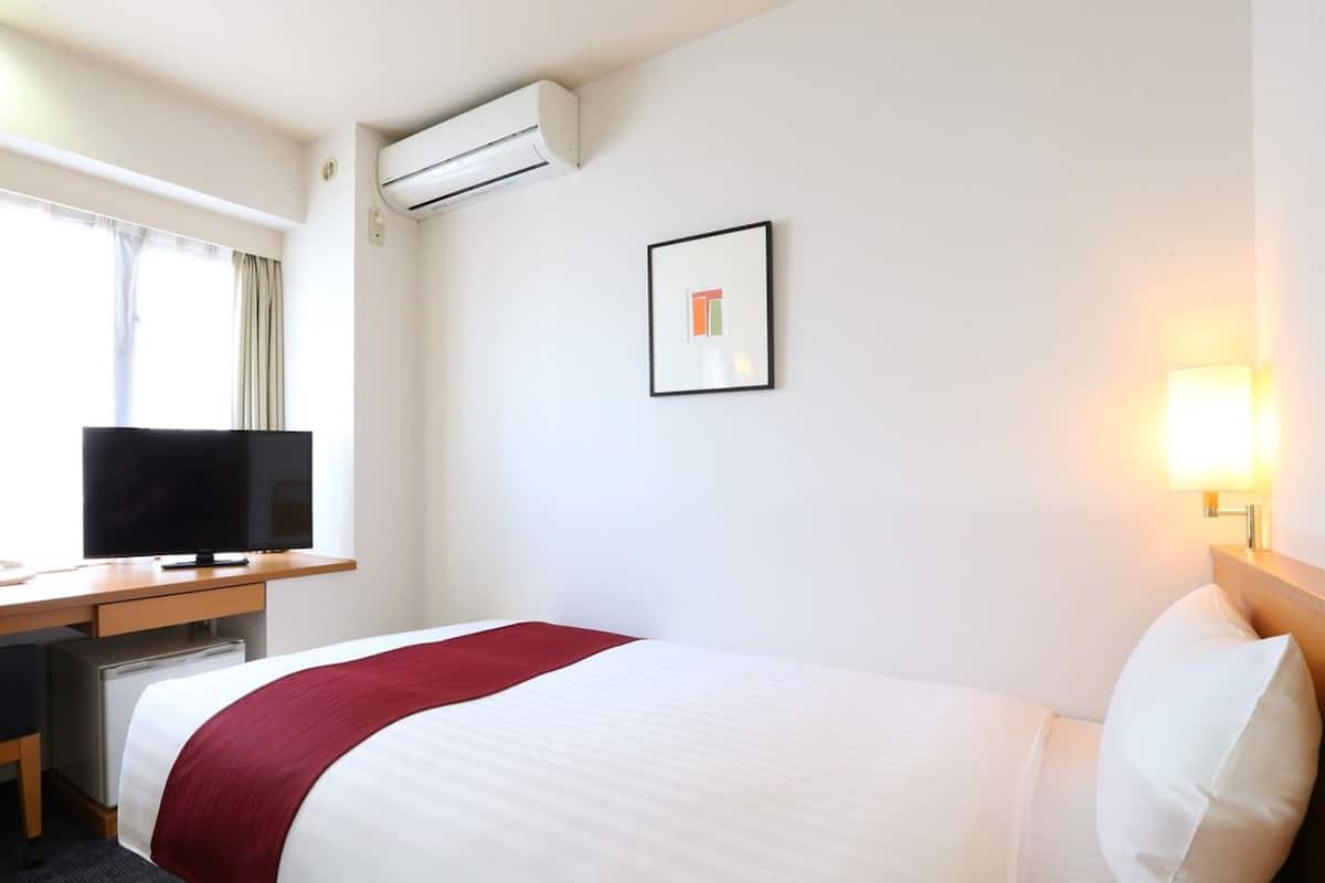 A hotel room with a bed, a desk with a television, an air conditioner, a framed picture, and a bedside lamp.