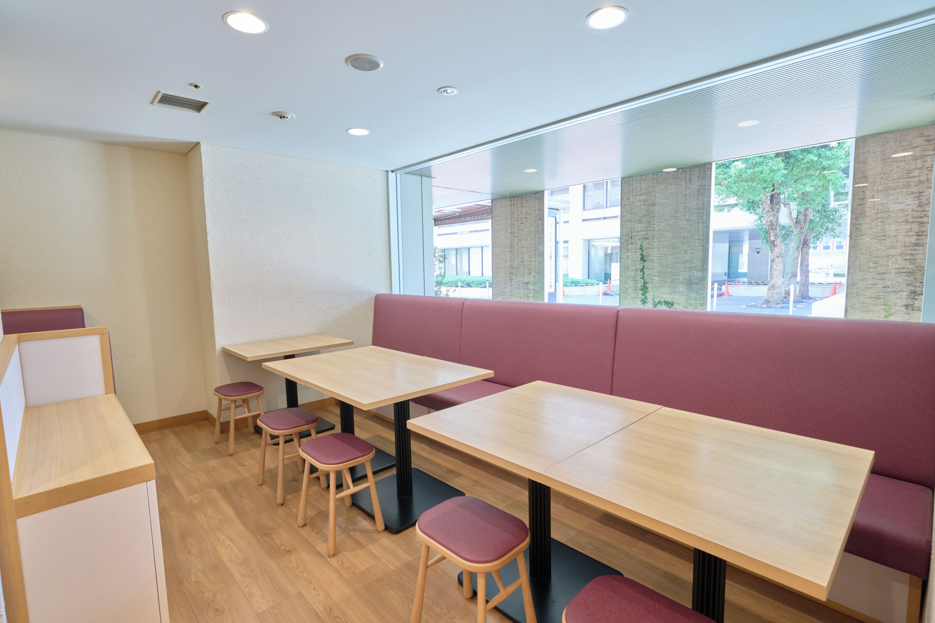 A restaurant interior with wooden tables and benches, a white wall, and a large window with a view of the outside.