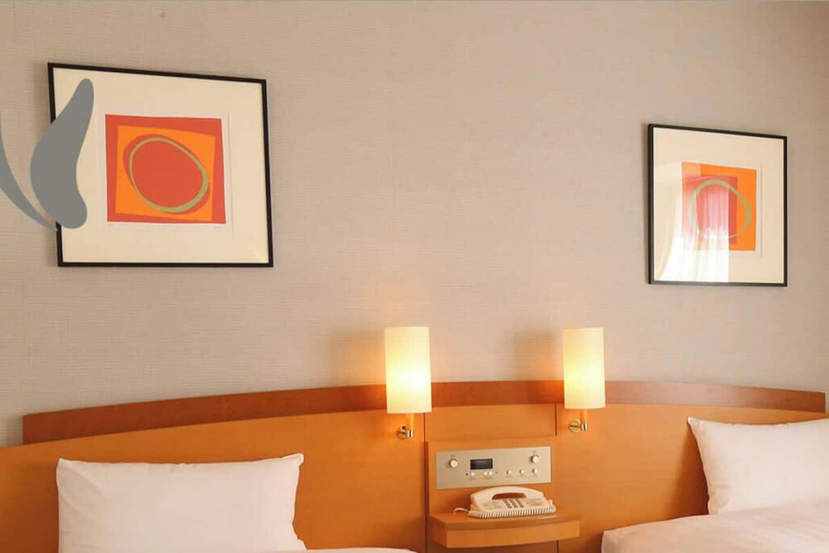 A hotel room with two beds, a headboard, bedside lamps, a telephone, and two framed pictures on the wall.