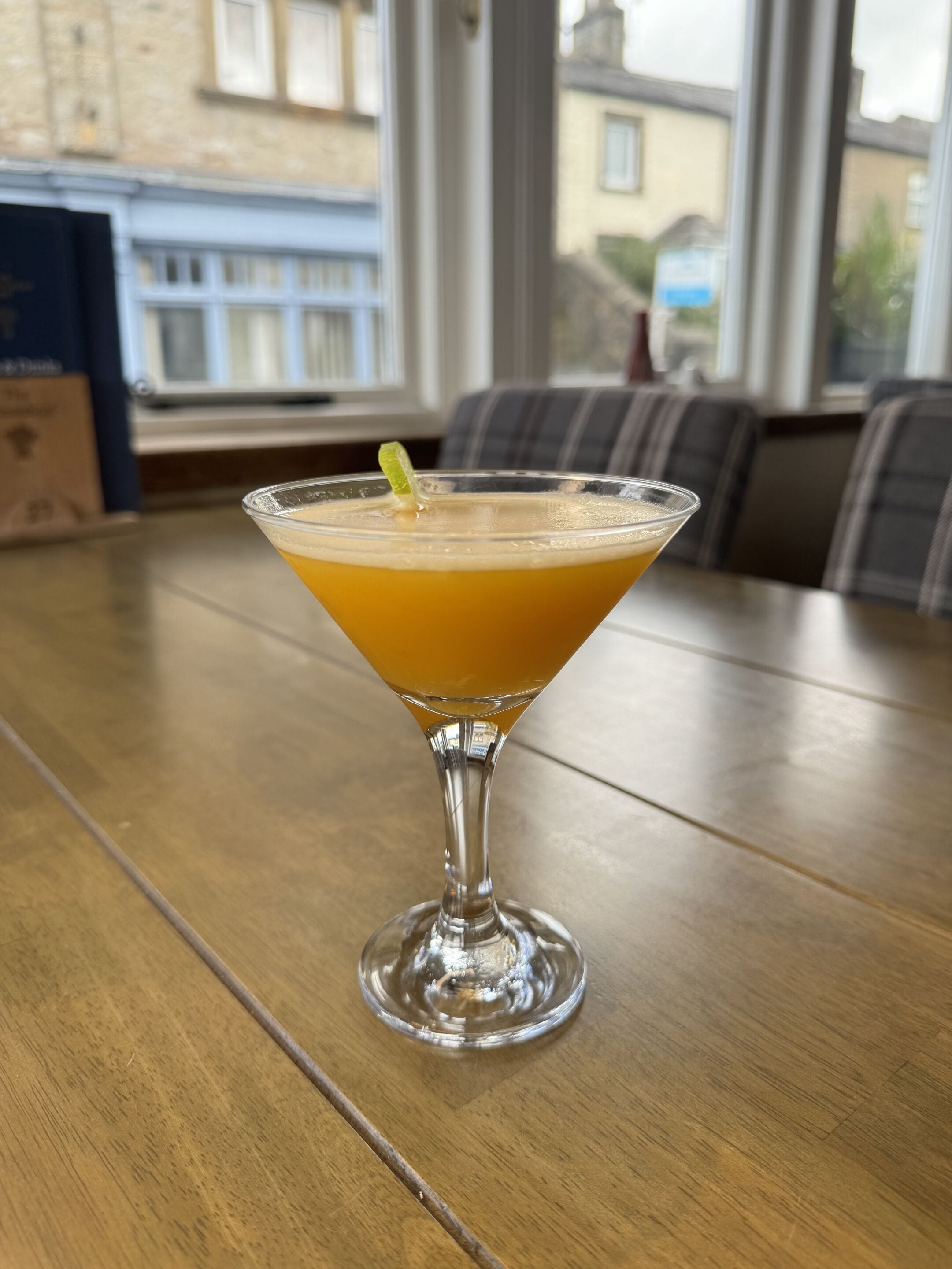A martini glass with a lime garnish on a wooden table in a room with a view of buildings through a window.