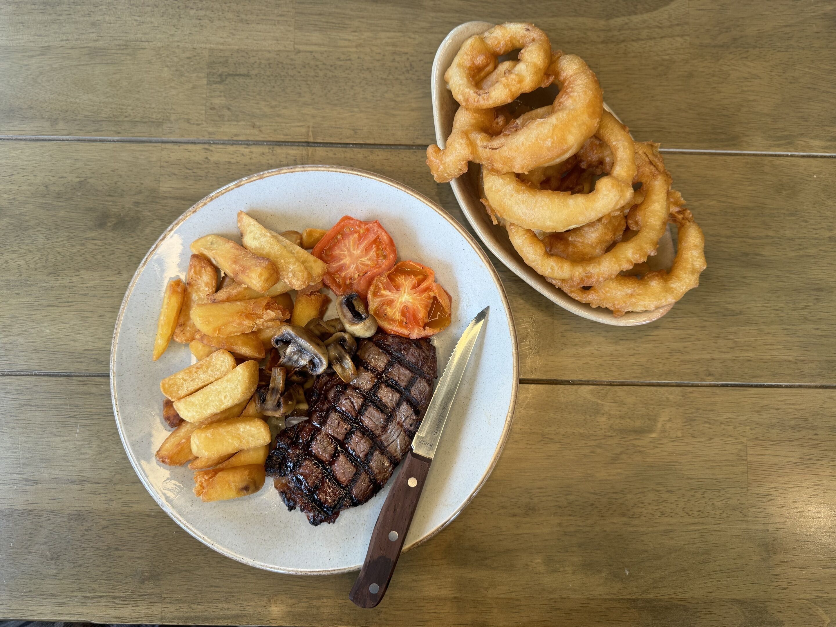 A plate with steak, fries, mushrooms, and tomatoes, and a separate plate with onion rings.