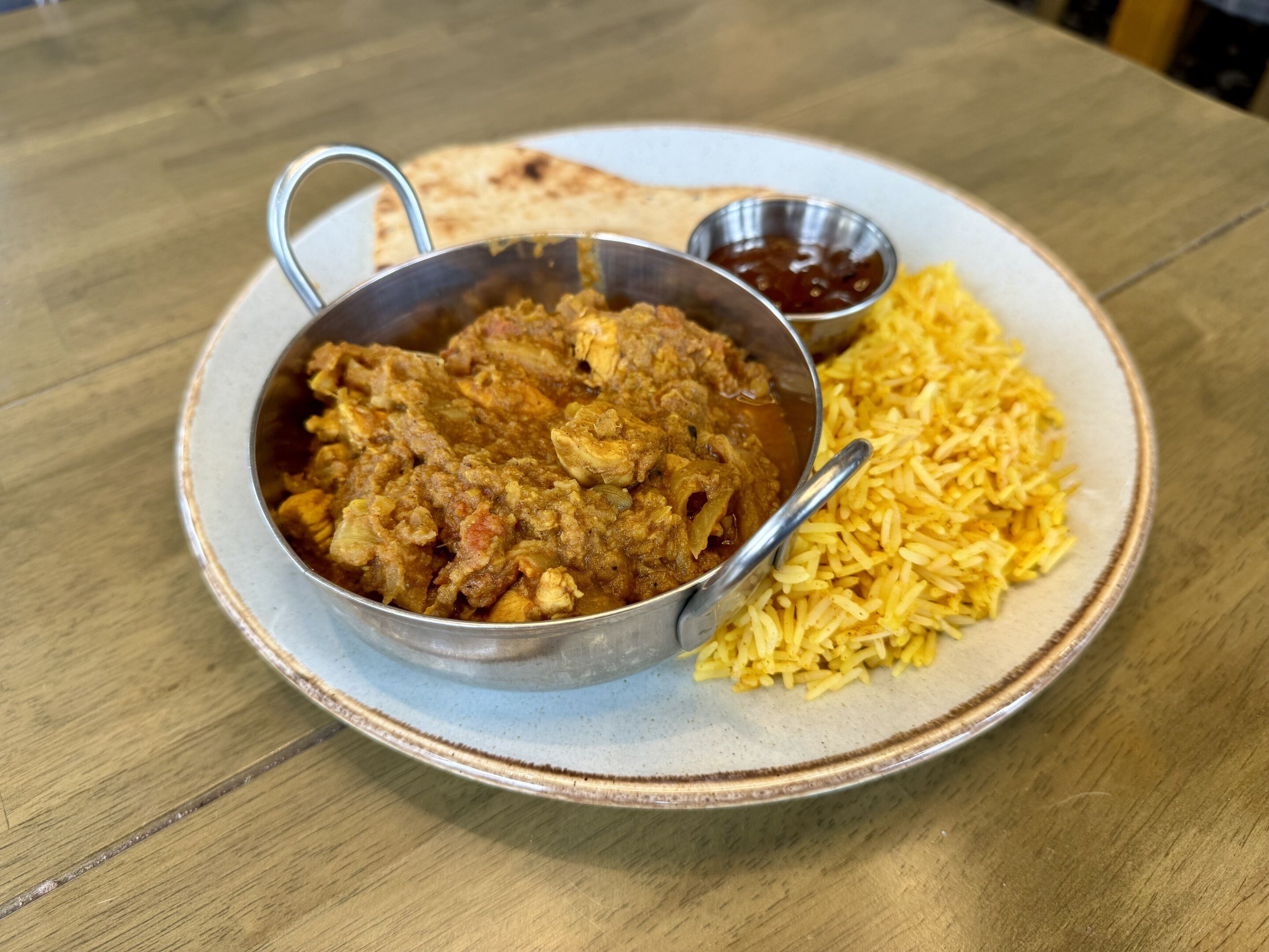 A plate with a curry dish and rice, accompanied by a piece of flatbread and a small bowl of sauce.