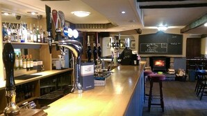 A bar area with a long counter, multiple taps, and a variety of bottles. There is a chalkboard menu and a fireplace in the background. - Wheatsheaf (Carnforth)