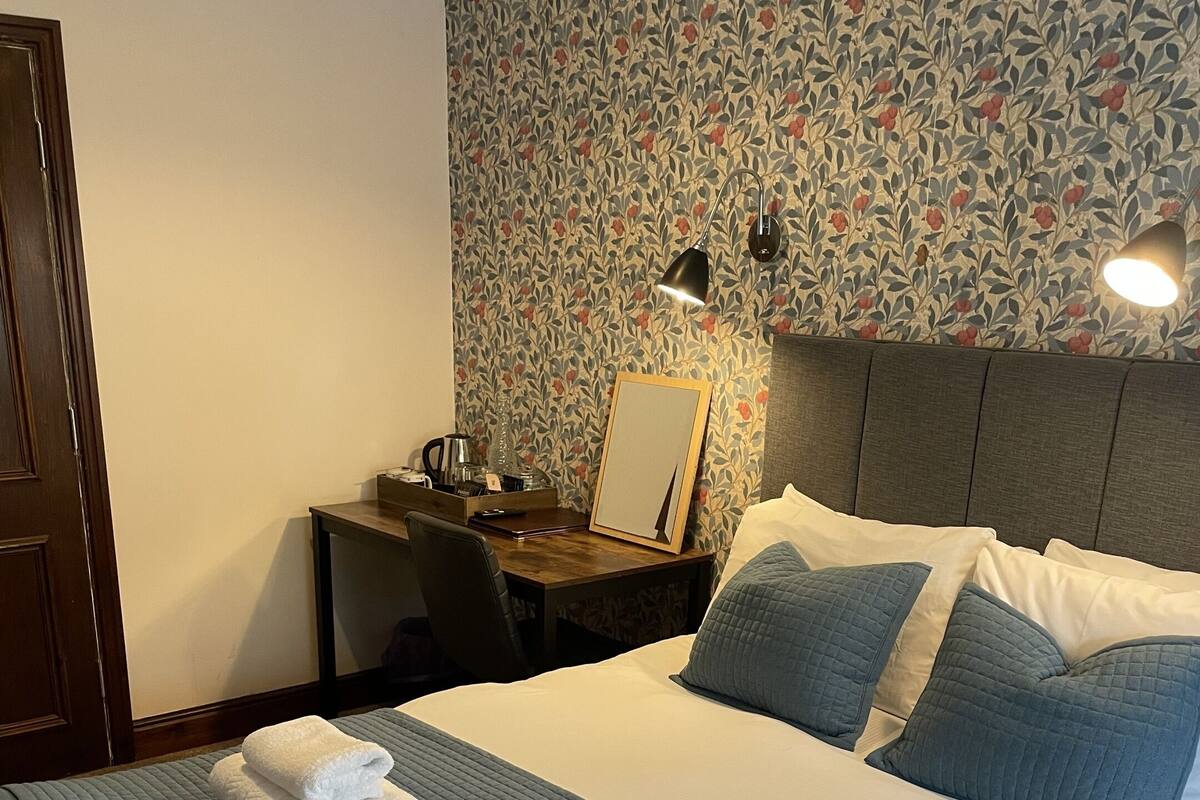A neatly made bed with a blue quilt and two folded white towels, a wooden nightstand with a lamp, a mirror, and a floral wallpaper pattern.