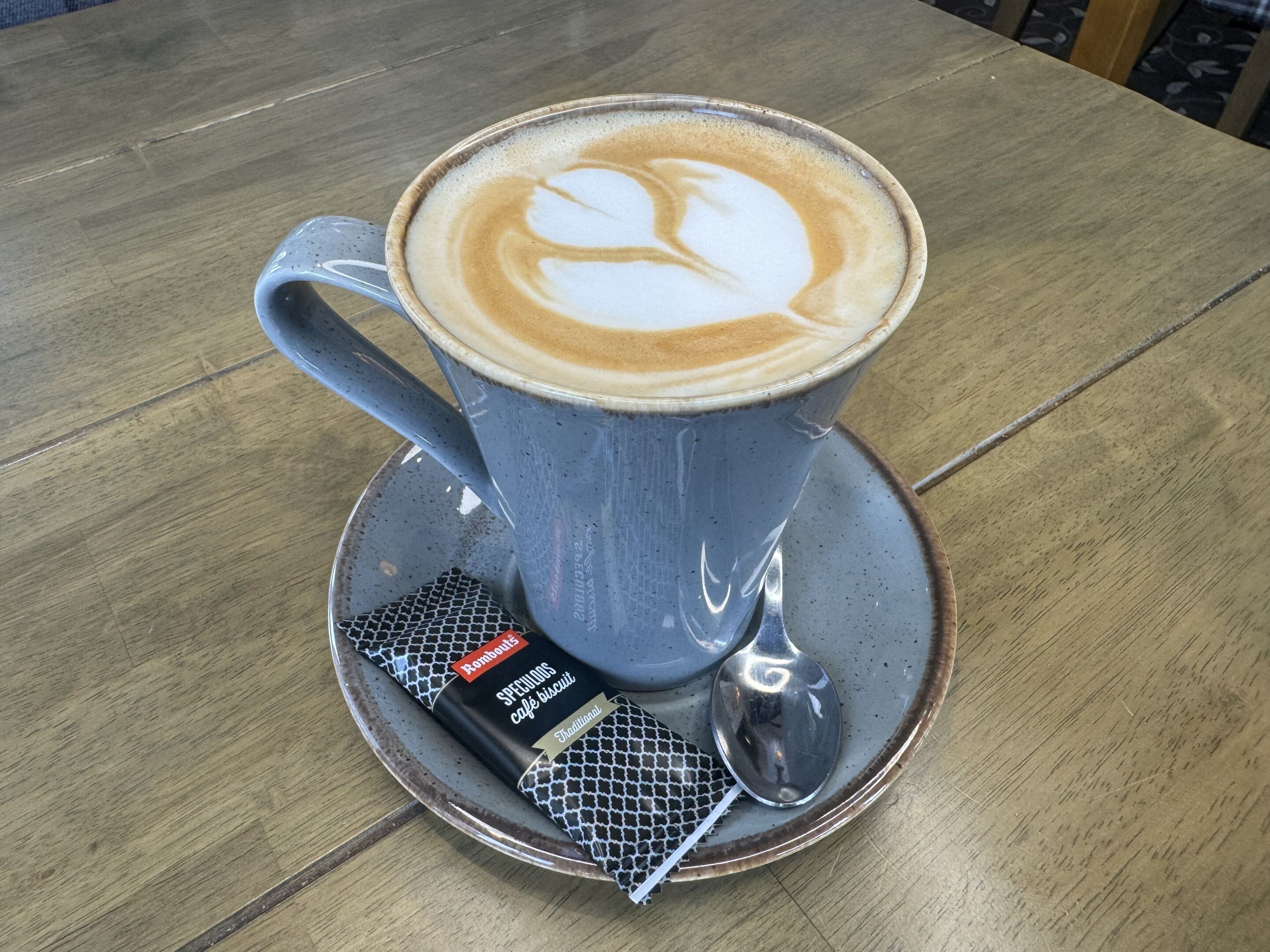 Une tasse de café décorée avec un motif en latte art, posée sur une soucoupe, accompagnée d’une cuillère et d’un paquet de biscuits au café.