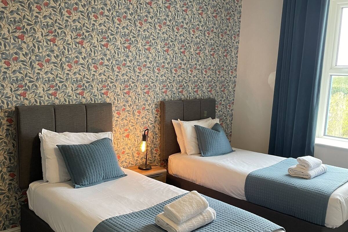A hotel room with two beds, a nightstand with a lamp, a window with curtains, and a patterned wallpaper.
