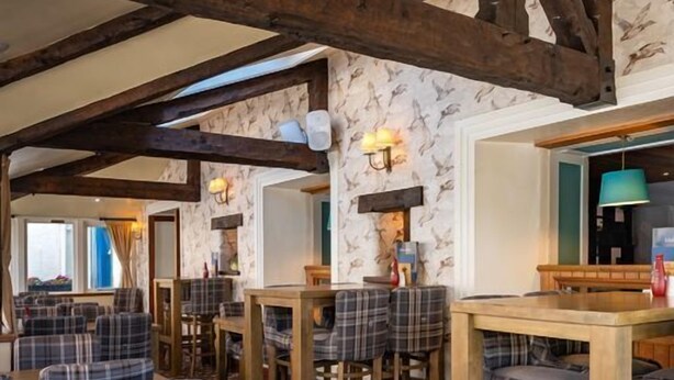 A restaurant interior with wooden tables and chairs, patterned wallpaper, and exposed wooden beams. - Wheatsheaf (Carnforth)