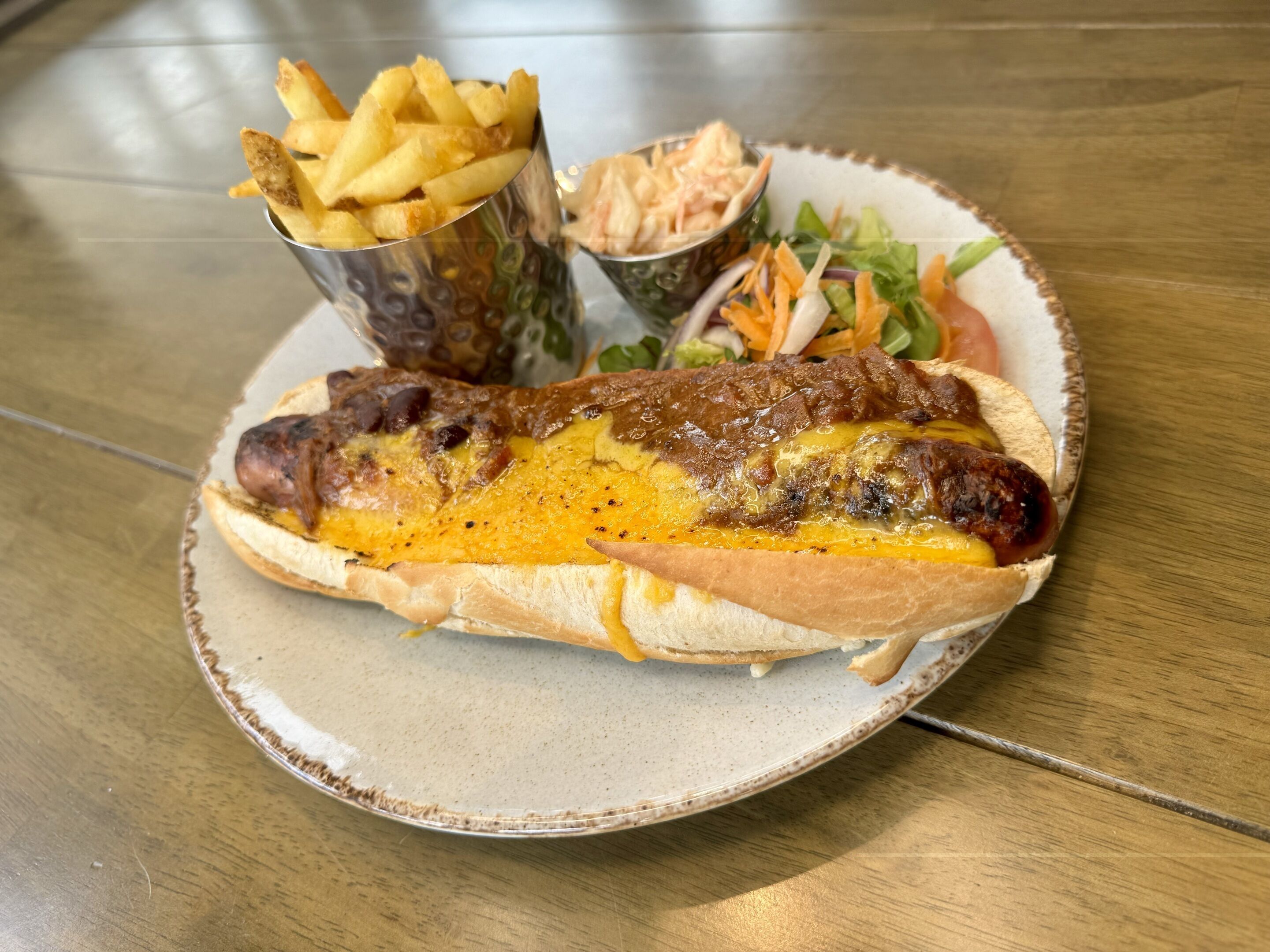 Ein Hotdog mit Käse und Senf auf einem Brötchen, dazu Pommes und ein kleiner Salat auf einem Teller.