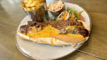 Un perro caliente con queso y mostaza en un pan, acompañado de papas fritas y una ensalada pequeña en un plato.